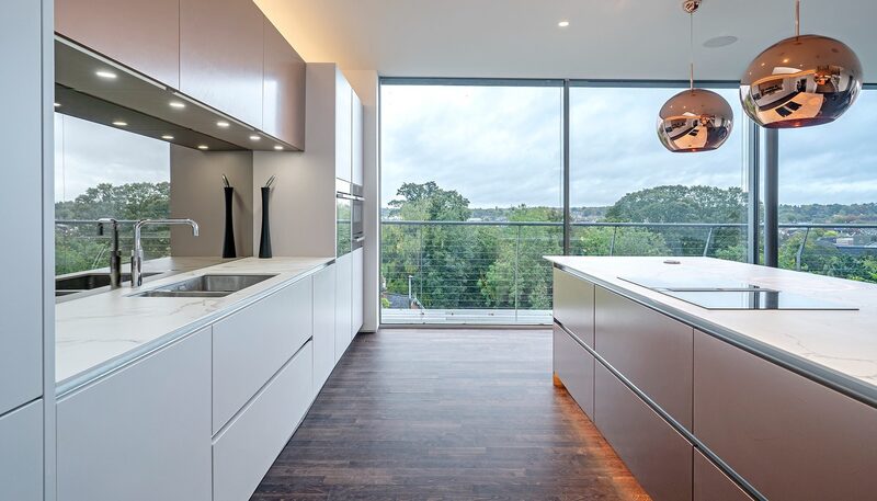 Porcelain vs quartz worktops: Watermark kitchen with pale worktops, copper pendant lights and full-height glazing overlooking trees.