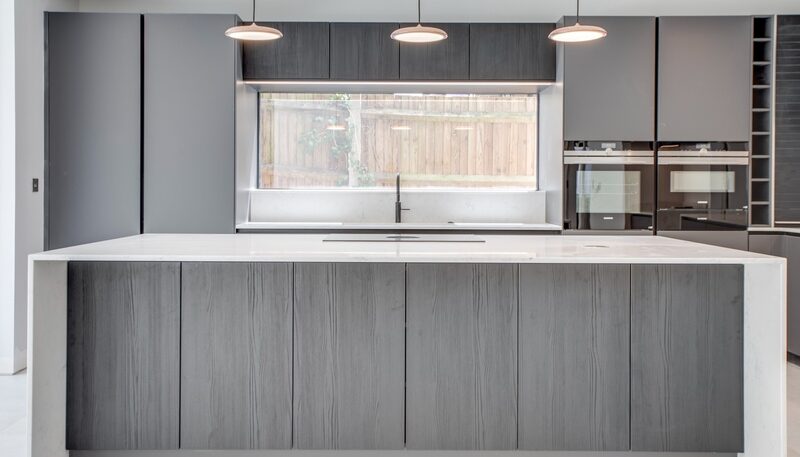 Porcelain vs quartz worktops: Watermark kitchen with grey cabinetry, a pale waterfall island and three pendant lights.